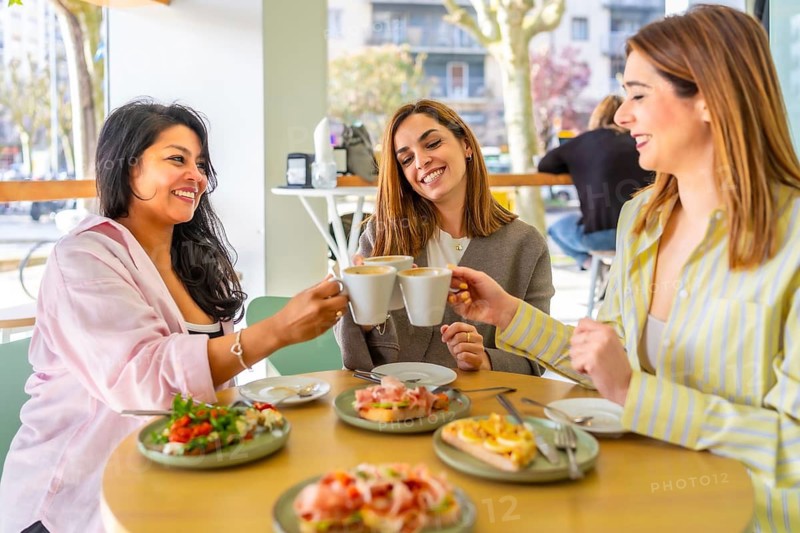 Three smiling women friends toast coffee cups at a bright cafe brunch, laughing and enjoying food together in a warm, casual, multi ethnic gathering of connection and joy
