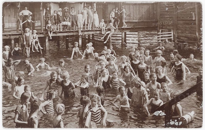 Bathing, swimwear, woman, men, children, leisure, fun, summer, many people in bathing suits playing and swimming in a water basin near wooden buildings, 1909