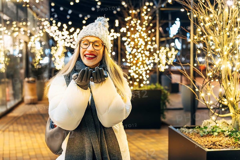 Young woman in winter clothing surrounded by festive lights. A woman wearing a white winter coat, gray knit hat with a pom-pom, dark scarf, gray gloves and glasses stands outdoors at night, smiling with her hands near her face. Warm white string lights wrap the nearby trees, creating a bright festive glow. Glass storefronts and the urban walkway add depth to the cozy Christmas and New Year atmosphere