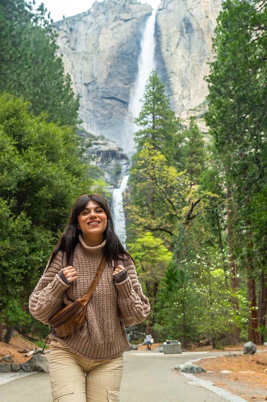 Young woman smiling while walking on a path, exploring the beautiful nature of yosemite national park with its towering lower yosemite fall and lush green trees