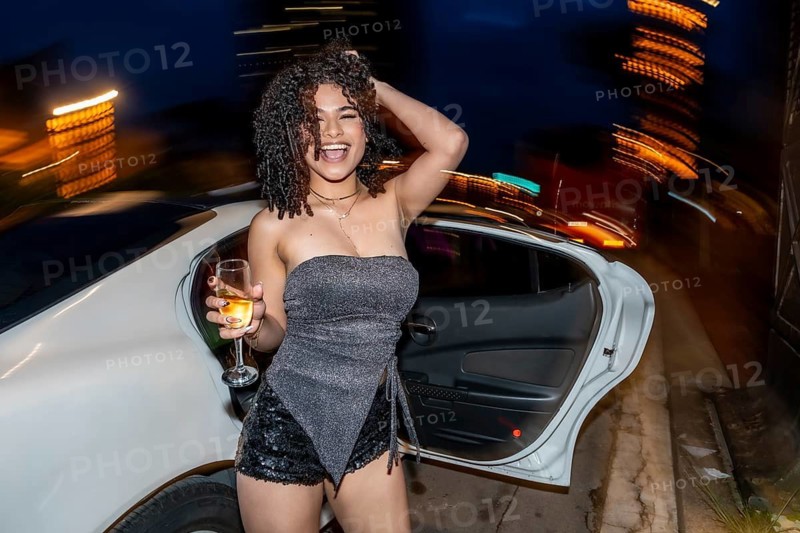 Young woman with curly hair laughing and holding a champagne glass, standing outside a luxury limousine at night, celebrating with city lights blurring in the background