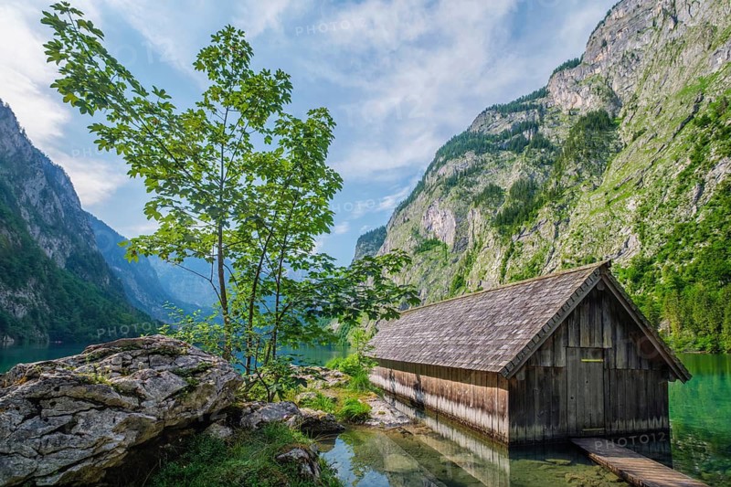 Wide angle view of the wooden boat shed at Obersee lake with a green tree on a rocky outcrop, Bavaria, Germany
