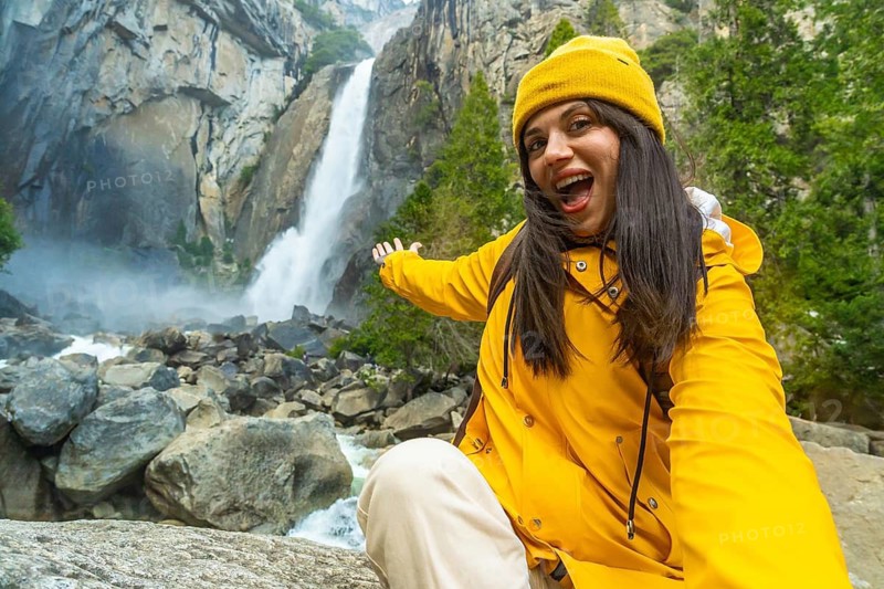 Woman in a yellow raincoat and hat smiling and pointing at lower yosemite fall, enjoying her hiking adventure in the granite valley and experiencing natural beauty