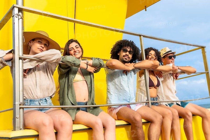Group of diverse friends leaning on a rail, smiling at the camera and celebrating friendship and summer vacation while sitting on a bright yellow lifeguard tower against a blue sky