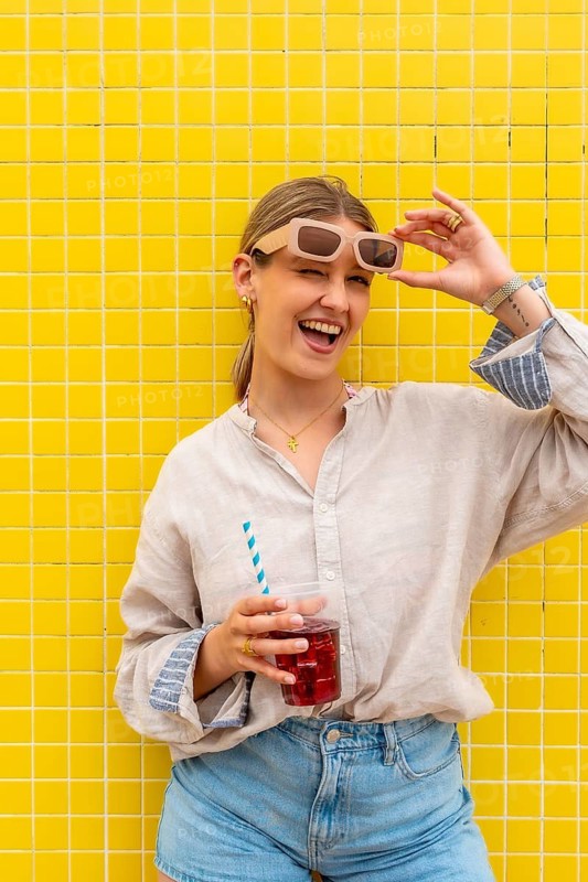 Happy young woman winking, laughing, and holding a refreshing iced drink, standing in front of a bright yellow tiled wall, enjoying a vibrant summer day with fashionable style