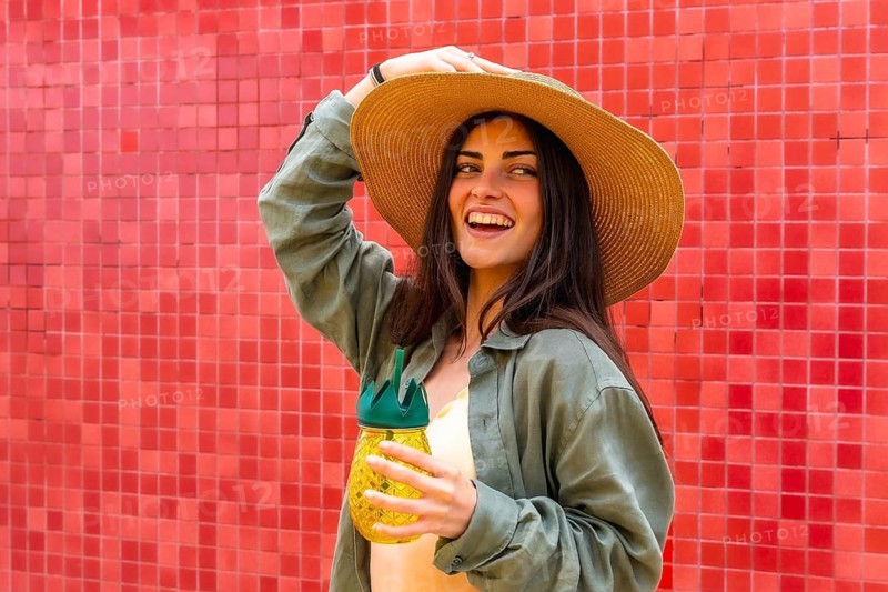Woman smiling brightly and posing in a straw hat, holding a refreshing pineapple shaped drink bottle against a vibrant red tiled wall, embodying summer joy and vacation vibes