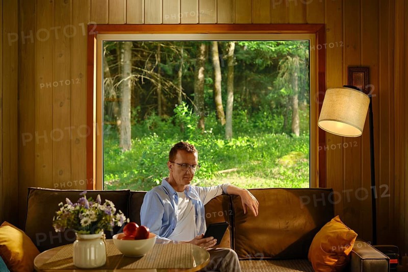 Middle-aged man hiding from heat in wooden cabin outside city sitting on couch and reading electronic book. Sunlight enters through large window. Outside, there are trees and green grass