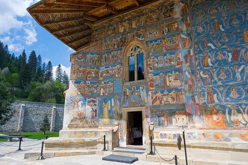 Church wall with colorful frescoes surrounded by trees under a blue sky, exterior view, Saint George Monastery, Voronet Monastery, Voronet Monastery, UNESCO World Heritage Site, Suceava County, Bukovina Region, Moldova, Romania