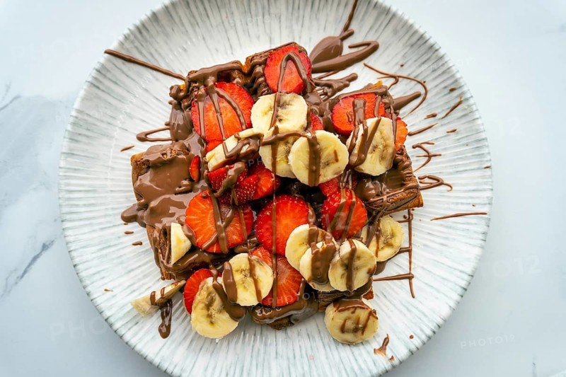 Close up top down view massive, high-calorie waffle topped with strawberries, bananas, and chocolate at an outdoor cafe. A delicious and indulgent big portion sweet treat