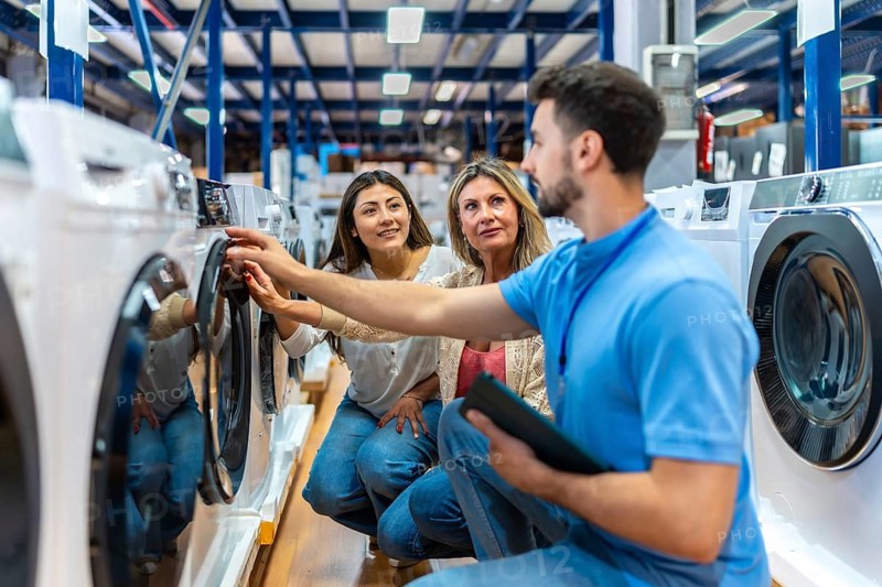 Sales associate helping a mother and daughter select a new washing machine at a home appliance store, discussing features and quality for their household needs
