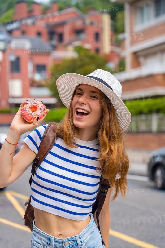 Young smiling woman wearing a hat and striped top holding a pink glazed donut with sprinkles, happy while exploring a city street on vacation