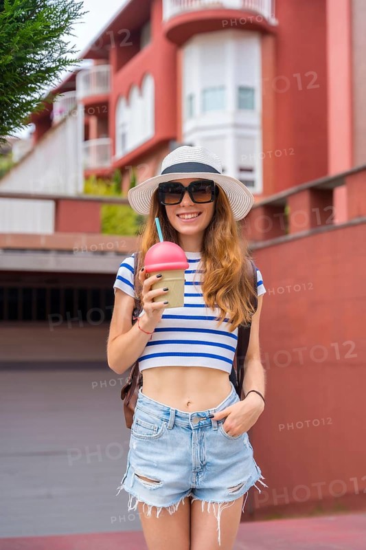 Young woman smiling while holding a refreshing beverage in a reusable cup, wearing a hat, sunglasses, striped top, and denim shorts, enjoying a bright summer day outdoors