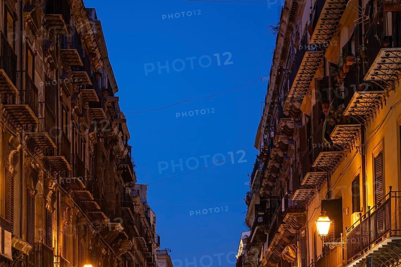 City street illuminated in the evening with tall old buildings and lantern light under clear sky, Palermo, Sicily, Italy