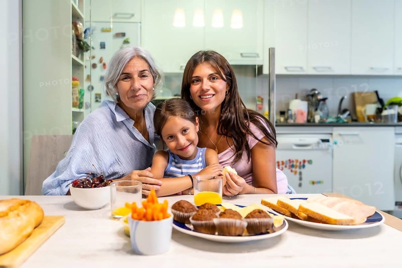 Multi generational women bonding, sharing a meal, and smiling at the camera at a kitchen table, symbolizing family love, connection, and daily life