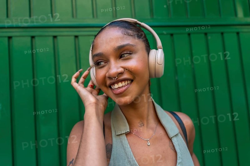 Young woman smiling while listening to music through wireless headphones, standing against a vibrant green wall, embracing technology and urban lifestyle