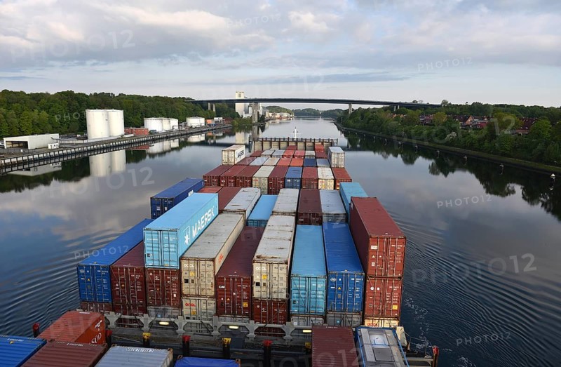 A container ship sails under a bridge on a quiet river, surrounded by trees, container ship sails in the Kiel Canal near Kiel-Holtenau, NOK, Kiel Canal, Kiel Canal, Schleswig-Holstein, Germany