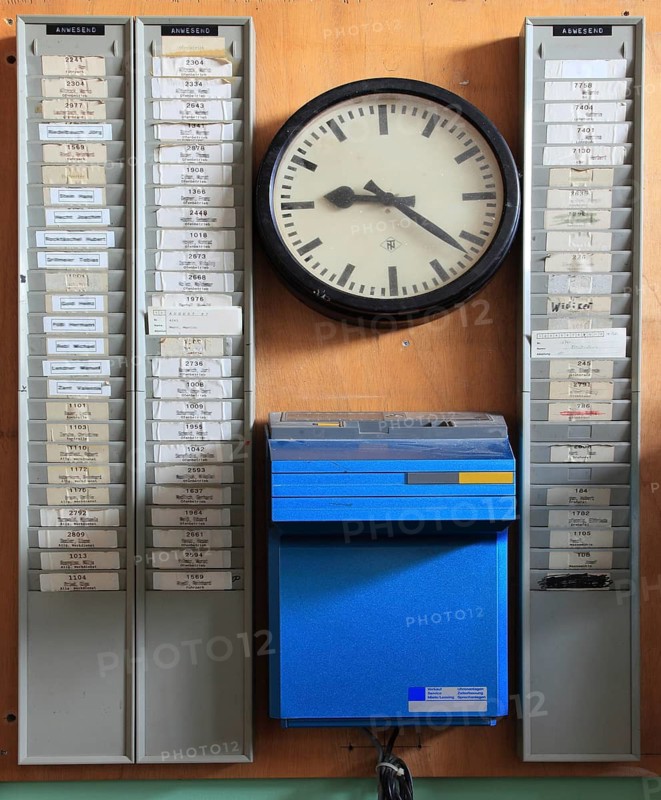 Office scene around 1972, lost place, abandoned location, historic time recording system from the 1950s, which contains a Solari DTC time clock, mechanical time clock, time clock for recording working time. The numbered stamp cards of the employees are stuck in the wall holders