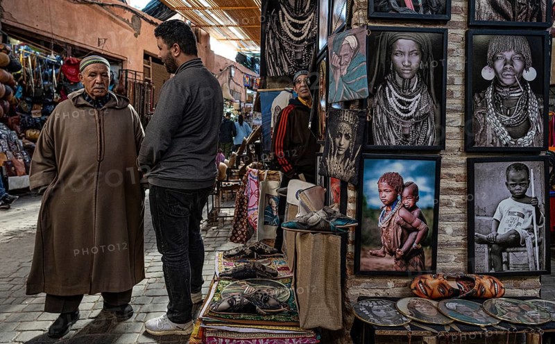 Two Moroccan men talk in the souk, bazaar next to a shop selling pictures, drawings of people in Africa, souk, bazaar, Marrakech, Maghreb, Morocco, North Africa