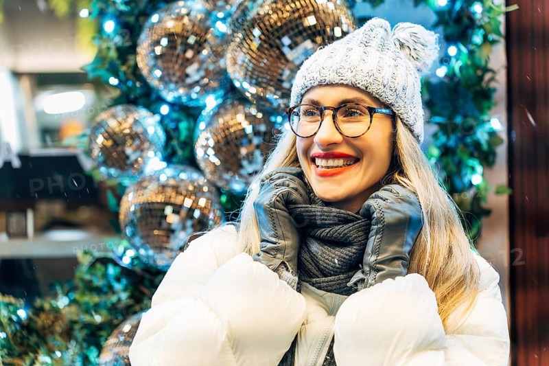 Young woman in winter clothing with festive decorations. A person in a white puffer jacket, gray knit hat with a pom-pom and glasses stands near large silver disco balls surrounded by greenery and blue-white lights. The reflective surfaces capture surrounding illumination, creating bright highlights and layered reflections. The festive winter lighting and decorative ornaments add a clear seasonal atmosphere to the scene