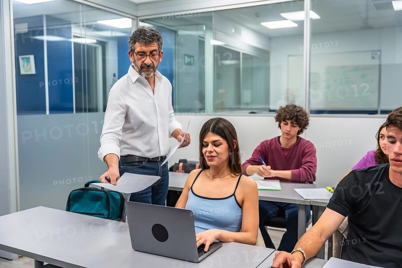 Professor interacting with diverse students in a bright school classroom, helping a young woman using a laptop for her studies while others are taking notes