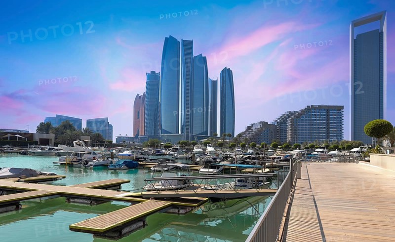 Iconic Abu Dhabi waterfront skyscrapers tower above a bustling marina filled with luxury yachts, viewed from a wooden boardwalk promenade at a colorful sunset, UAE