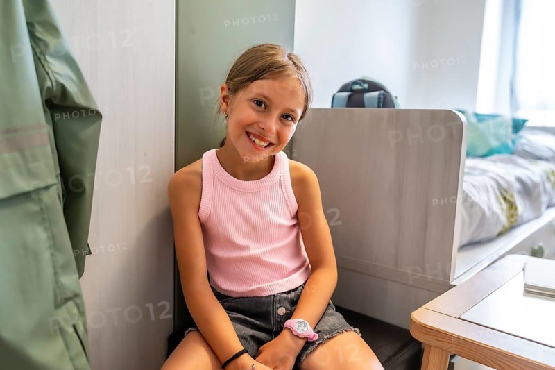 Young caucasian girl smiles happily while sitting on floor inside her bright modern bedroom with furniture