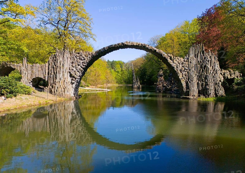 Arcaded stone bridge over a clear pond surrounded by lush green trees under a blue sky, aerial view, Rakotz Bridge, Devil's Bridge, Rakotzsee, arched bridge, reflection, basalt organ, Kromlau, Görlitz district, Saxony, Germany