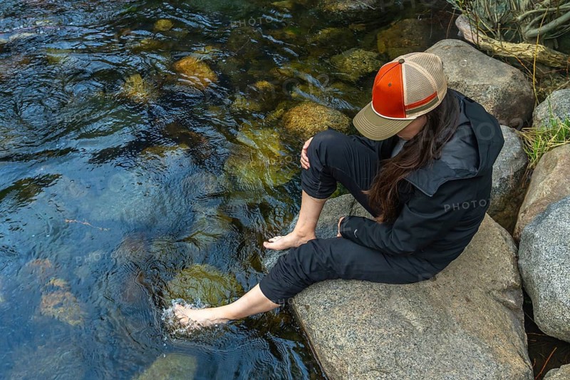 Woman dipping bare feet into a shallow, clear mountain river, enjoying the refreshing cool water while sitting comfortably on a large river rock in a peaceful natural environment