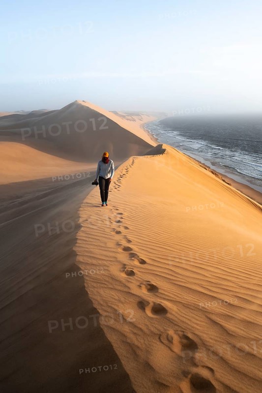 Young woman with camera, sand dunes with wave pattern and sea in evening light, wind blowing sand across the dunes, Atlantic coast and sandy desert with sea of dunes, impressive desert landscape at sunset, Sandwich Harbour, Namib Naukluft Park, Namib Desert, Namibia