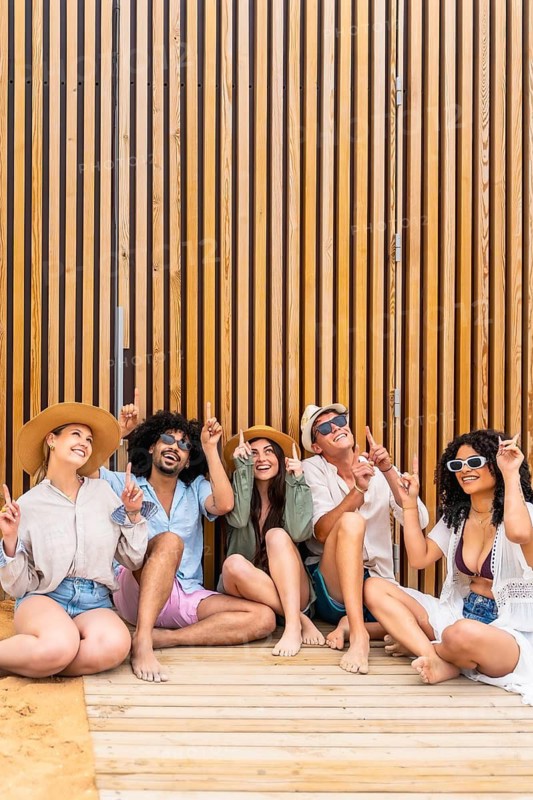 Diverse group of young adult friends sitting on a wooden deck next to a modern slatted wall, smiling and looking up, celebrating summer vacation and togetherness