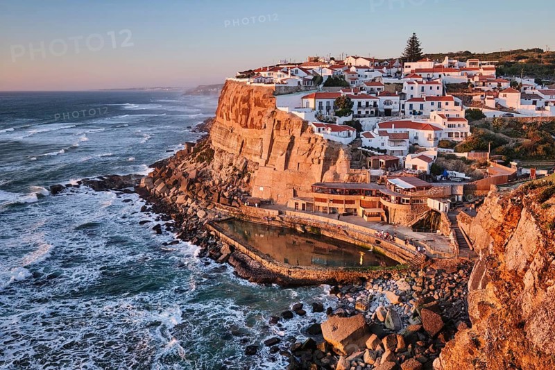 Breathtaking view of Azenhas do Mar, a charming seaside village perched on cliffs by the Atlantic ocean. The warm colors of sunset highlight the picturesque homes and rocky shoreline. Portugal