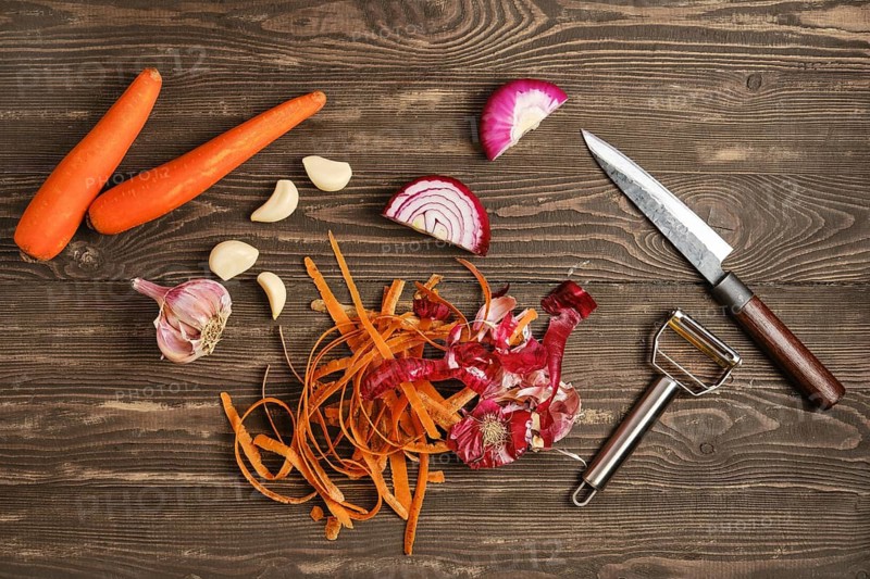 Two carrots, several garlic cloves, and pieces of red onion are laid out on a wooden table. A knife and vegetable peeler sit nearby, showing the process of food prep for cooking