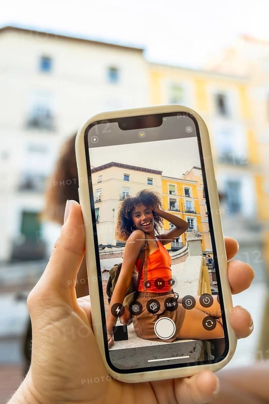 Hand holding a mobile phone recording a happy young woman with curly hair smiling for the camera, posing outdoors in a european city street on a sunny day