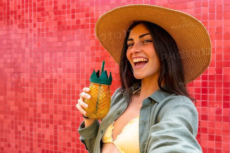 Woman in a straw hat and bikini top laughing while holding a tropical pineapple drink, taking a selfie against a vibrant red tiled wall, embodying summer vacation happiness