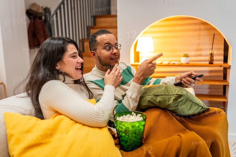 Happy diverse couple relaxing on a couch, holding popcorn, pointing while enjoying an entertaining movie night together in their cozy living room at home