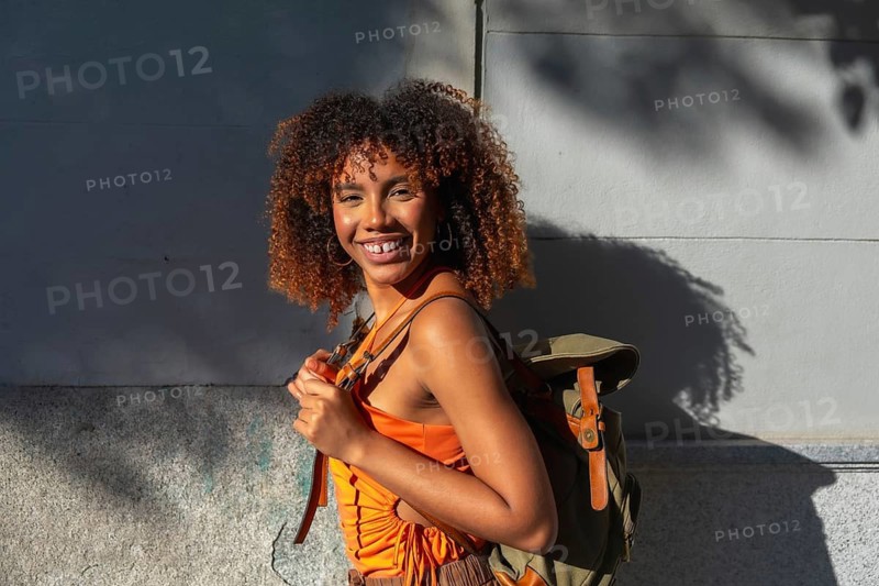 Young woman with natural curly afro hair and backpack walking in sunlit city streets, smiling cheerfully, casting warm shadows on urban walls, carefree and confident