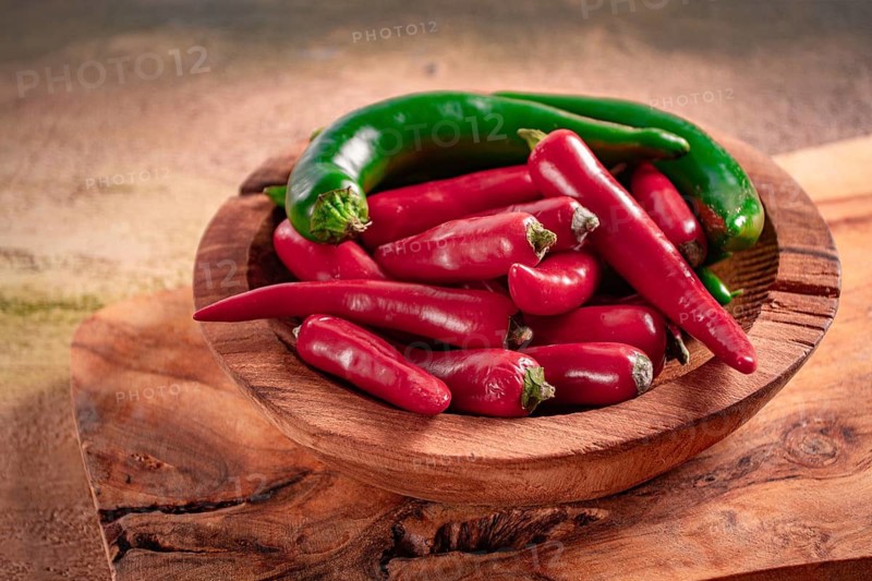 Bright green and red peppers are neatly placed in a round wooden bowl on a wooden board. The board has a natural finish, showing the grain of the wood