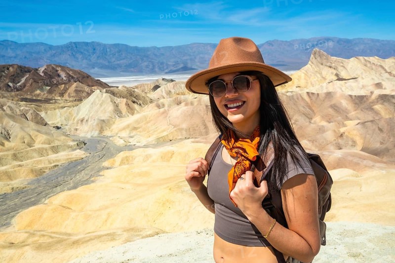 Young woman smiling, wearing a hat and sunglasses, holding a backpack while exploring the unique badlands landscape and vibrant geological formations of death valley national park