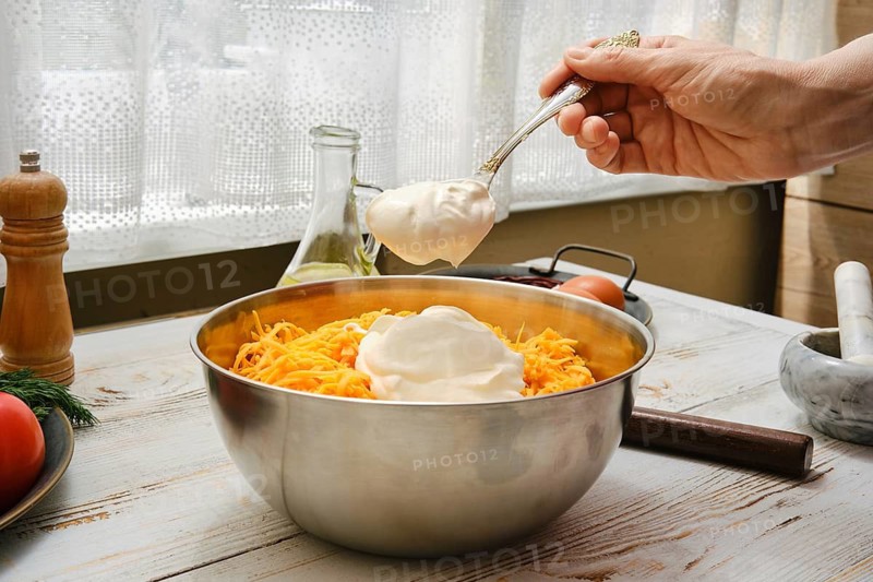 A person adds a spoonful of sour cream to a bowl of shredded cheese and chicken meat in a kitchen. The scene has various kitchen items on a table and sunlight coming through the window