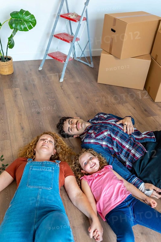 Happy family of three, parents and daughter, lying on the floor surrounded by moving boxes and a ladder, experiencing joy and relief in their new home