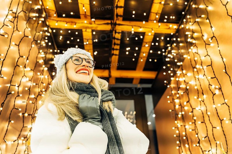 Young woman in winter clothing looking up under festive lights. A woman wearing a white winter coat, gray knit hat, scarf and gloves stands beneath a canopy of warm white string lights hanging from wooden beams. The dense illumination creates a bright festive glow around her, highlighting winter textures and the cozy Christmas atmosphere of a nighttime seasonal display