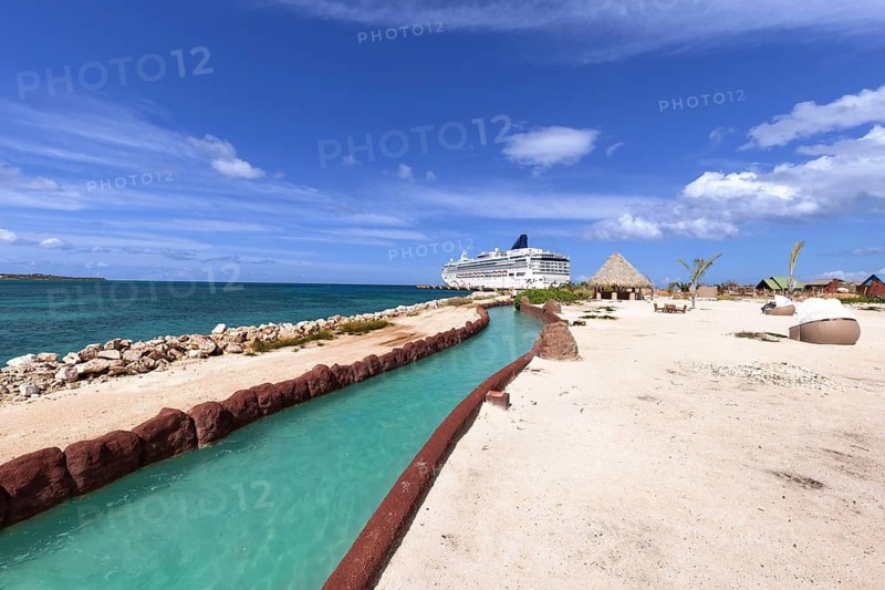 Cruise ship docked at Cabo Rojo port, Puerto Plata, Dominican Republic, featuring a winding turquoise lagoon canal, white sand beach, thatched huts, and vivid blue Caribbean sky
