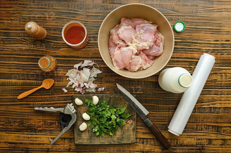 Chicken thighs are in bowl surrounded by spices, herbs, garlic, and yogurt. A knife and cutting board are also present. This scene shows cooking setup for meal preparation at home