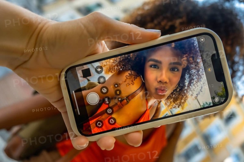 Hand holding smartphone showing woman with curly hair puckering for a selfie on the front camera, playful influencer moment captured on screen in casual orange top during daytime outdoors