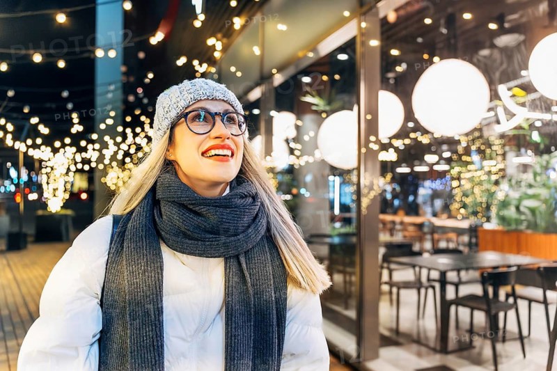 Young woman enjoying festive lights in winter evening. A woman wearing a light gray knit hat, glasses, dark scarf, gray gloves and a white puffy jacket stands outdoors at night beside a glass-front café glowing with warm round lights. Decorative string lights wrap nearby trees and poles, creating a bright festive holiday atmosphere. The mix of winter attire, illuminated urban surroundings and cozy seasonal lighting conveys a strong Christmas and New Year mood