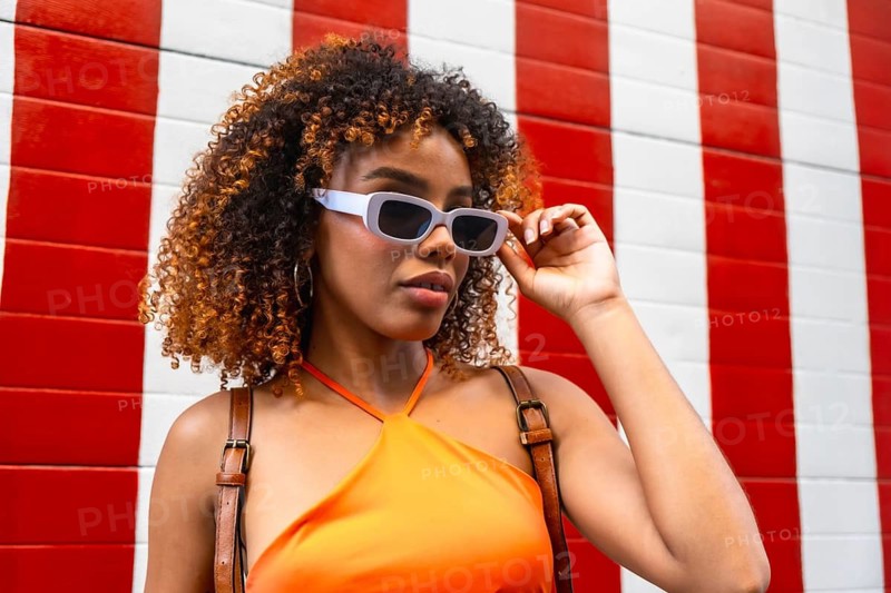 Young woman with natural curly hair and an orange top, wearing cool sunglasses and posing confidently in front of a modern red and white striped background