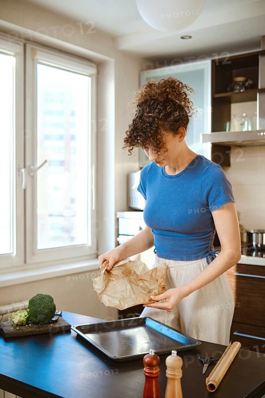 Woman is handling parchment paper near a baking tray. Natural light brightens the space