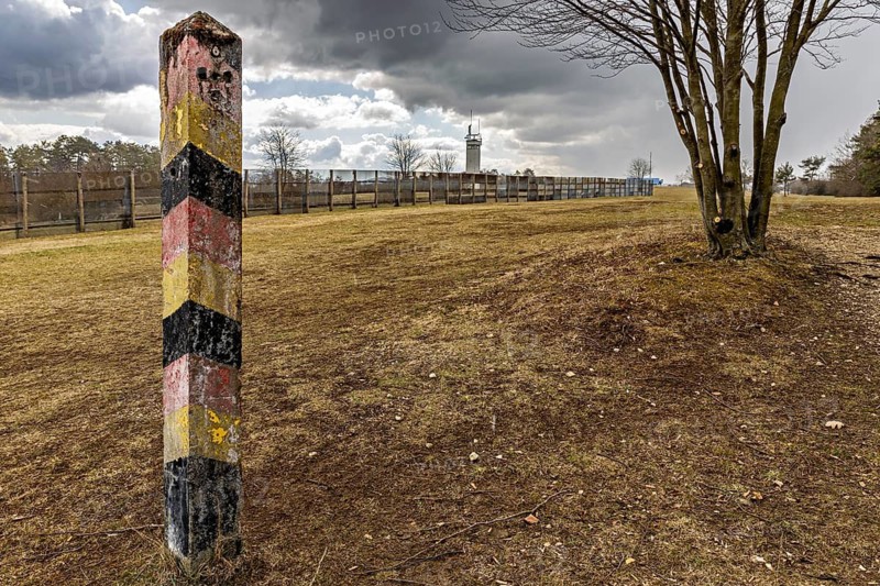 A colorful landmark stands in a meadow with a tree and a distant watchtower in cloudy skies, the Point Alpha memorial between Rasdorf and Geisa in the Rhön