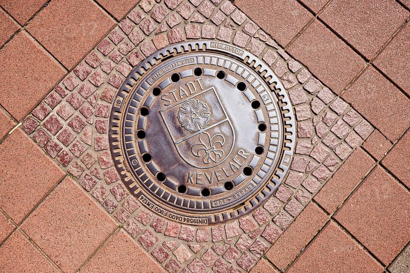 Manhole cover, city coat of arms, city of Kevelaer, footpath made of concrete paving stones and paving stones, sunny, Kevelaer, Kleve district, North Rhine-Westphalia, Germany