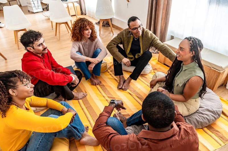 Diverse adults sitting in a circle on the floor, sharing thoughts and listening to each other during a supportive group session focused on mental health and community building